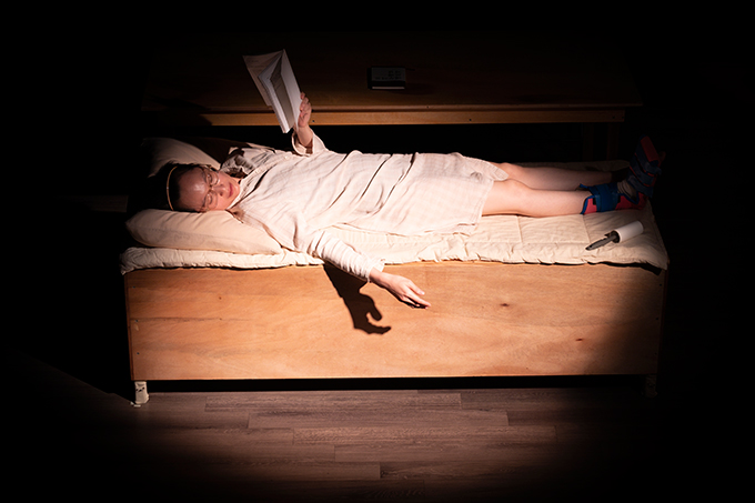 <애 개 아파트>의 공연 사진. 침대 위에 누워 한 손으로 책을 들고 있는 신소우주의 모습이다. 그는 다른 손을 침대 밖으로 늘어뜨린 채 바닥 쪽을 바라보고 있다.