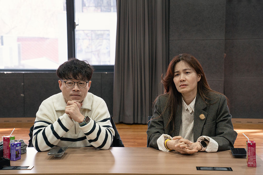 연출가 김태현과 배우 김순덕이 나란히 앉아 이야기를 경청하고 있다. 김태현은 턱 아래에, 김순덕은 책상 위에 붙잡은 양손을 모으고 있다.