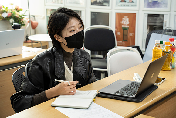 좌담 진행자 김연재. 짧은 커트 머리에 검은색 재킷을 입고 검은색 마스크를 쓰고 있다. 오른손에 볼펜을 들고 있고, 책상 위에는 메모를 할 수 있는 다이어리와 노트북이 펼쳐져 있다. 