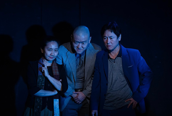 <맹>의 공연 사진이다. 파란 색 옅은 조명 아래 세 명의 배우들이 어깨를 맞대고 심각한 표정으로 나란히 서 있다. 
