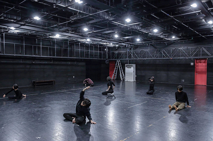 검은 벽과 바닥으로 된 연습 공간에서 여섯 명의 배우가 각자 바닥에 앉아 자유롭게 몸을 풀고 있다.