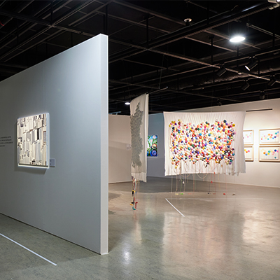 지금 보시는 공간은 현대적인 느낌의 실내 전시장입니다. 천장이 높고 넓은 공간에, 전반적으로 어두운 조명이 깔려 있으며, 벽과 바닥은 흰색과 회색 계열로 차분한 분위기를 자아냅니다. 천장에서 바닥까지 길게 늘어진 여러 장 매달려 있습니다. 왼쪽 벽면에는 전시 설명이 적힌 텍스트와 함께, 파란 조명이 들어오는 디지털 화면이 보입니다. 벽면에는 여러 색의 선들이 그어져 있고, 그 옆으로 텍스트가 배열되어 있어 전시의 주제나 개념을 소개하는 것으로 보입니다. 오른쪽으로는 낮은 조명 아래, 여러 개의 작품이 벽에 나란히 걸려 있습니다. 작품은 각각의 크기와 구성이 조금씩 다릅니다. 기둥과 벽 사이에 충분한 공간이 있어 관람객이 여유롭게 움직이며 감상할 수 있도록 구성되어 있습니다. 바닥은 반사되는 재질로 되어 있어 천장과 조명이 은은하게 비쳐 보이며, 전시 전체에 고요하고 집중된 분위기를 연출합니다. 이 전시 공간은 정적인 분위기 속에서 관람자가 하나하나의 작품에 천천히 몰입할 수 있도록 설계되어 있습니다.