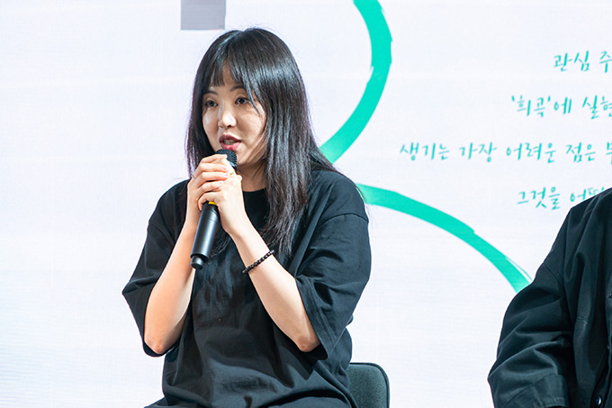 작가 신효진. 앞머리가 있고, 층을 낸 머리가 어깨 아래로 내려온다. 검은 반소매 티셔츠를 입고 있으며, 왼쪽 팔목에 검은 구슬 팔찌가 보인다. 마이크를 잡고 이야기하는 중이다.