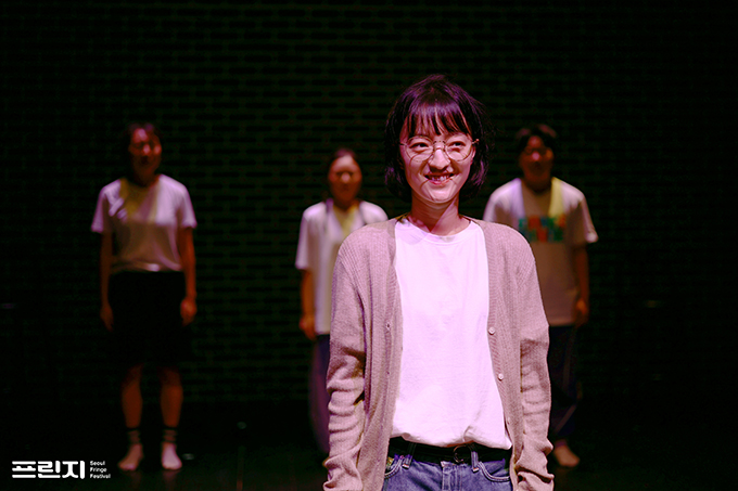 <홀리섹스데이> 공연 사진. 안경을 쓴 단발의 슬아가 정면을 보며 장난스럽게 웃고 있다. 그는 흰 티셔츠와 청바지, 베이지색 가디건을 입었다. 
            슬아의 뒤로는, 정자세로 서서 그녀를 바라보는 세 사람의 모습이 흐릿하게 보인다. 