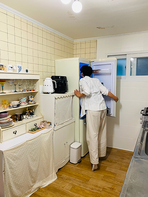 <맡는 경계>의 전시 사진. 베이지색 타일 벽이 눈에 띄는 가정집의 풍경이다. 
            위아래로 하얀색과 옅은 베이지색의 옷을 입은 한 사람이 냉장고의 문을 열고, 상체를 앞으로 숙여 냉장고 안 냄새를 맡는 뒷모습이 보인다. 
            타일 벽면은 찻잔과 고블린 잔, 접시 등이 수납된 장식장과, 커피포트와 에어프라이어가 놓여있는 전자레인지 수납장, 냉장고로 채워져 있다. 
            모두 하얀 톤의 가구들이다. 맞은 편에는 가스레인지와 싱크대의 일부가 보인다.