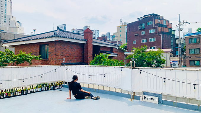 <맡는 경계>의 전시 연계 퍼포먼스 사진. 주변의 집들보다 층수가 비교적 낮은 주택 건물의 옥상에 검은색 상하의를 입은 퍼포머가 앉아있는 뒷모습이 보인다. 
            퍼포머는 두 다리를 모아 오른편으로 가볍게 뻗고 왼손은 땅을 짚은 채, 오른손은 다리 위에 가볍게 얹은 자세다. 
            앉아 있는 퍼포머보다 조금 높은 철제 난간은 흰 천으로 막혀 있어 난간 사이로 풍경이 보이지는 않는다. 
            난간에 길게 늘어뜨린 전선 사이사이 알전구가 달려 있고, 오른편 천 아래 ‘몸소리말조아라’의 로고가 쓰인 작은 간판이 보인다. 왼편 난간에는 빈 술병이 빼곡히 놓여 있다.