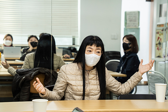 참여자 조아라. 허리까지 오는 생머리에 눈썹 위로는 짧은 앞머리가 내려와 있다. 베이지색 재킷을 입었고 하얀 마스크를 썼으며, 양팔을 벌린 채 이야기를 하고 있다.