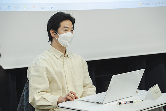 작가 이민규. 옅은 노란색 셔츠 차림이다. 그의 뒤편으로 프로젝터 화면의 하단이 조금 보인다. 