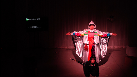 <안타 오이디푸스: hit or run>의 공연 사진. 흰 고깔 모자와 흰 망토를 입은 사람이 붉은 조명 아래 양팔을 좌우로 벌리고 무표정하게 서 있다. 그가 입은 옷은 파랑, 빨강, 노랑, 초록색이 양팔과 가슴에 T자로 나열되어 무당을 연상케 한다. 그 앞에는 검은 야구잠바를 입은 여성이 무릎을 꿇고 두 손을 짚은 채 고개를 들고 있다. 사진 왼편 스크린에는 ‘웅장하고 무거운 효과음’이라는 문자 해설과 T의 대사가 보인다.  