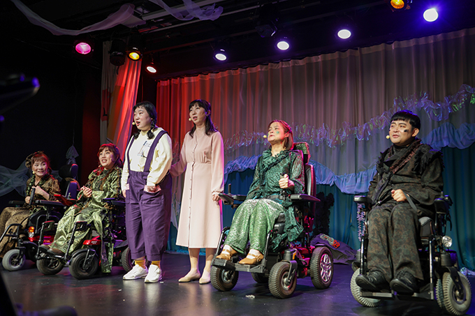 <숨 쉬는 바닷말> 공연 사진. 톳, 미역, 소녀(아자), 엄마, 파래, 김 역할의 배우들이 일렬로 나란히 자리 잡고 노래를 부르고 있다.
톳, 미역, 파래, 김 역할의 배우들은 모두 전동휠체어를 타고 있으며, 옷깃과 소매 등에 레이스 프릴이 달린 초록색과 검은색 계열의 의상을 입었다.
아자는 흰색 티셔츠에 보라색 멜빵 바지를 입었고, 엄마는 옅은 분홍색 원피스를 입었으며 아자의 팔짱을 끼고 있다.
배우들로 가득 찬 무대의 뒷벽에는 흰색과 초록색 커튼이 레이어를 만들고 있으며, 여러 색깔의 화려한 조명이 이들을 비추고 있다.