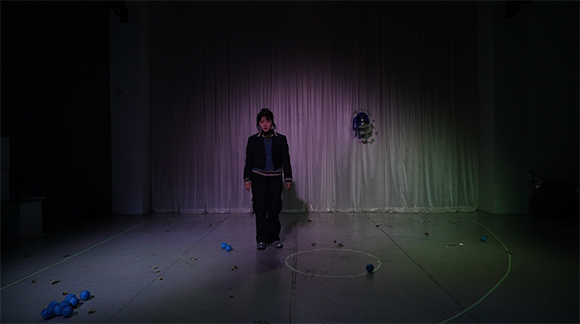 <안타 오이디푸스: hit or run>의 공연 사진. 화이트 박스 무대의 뒤편에는 두 개의 기둥 사이로 흰 커튼이 있다. 바닥에는 흰 선으로 야구장과 같은 부채꼴 모양이 그려져 있고, 그 가운데 마운드를 연상시키는 둥근 원이 그려져 있다. 무대 가운데에는 검은 야구점퍼와 검은 바지를 입고 단발 머리를 반묶음한 여성이 바닥을 보며 서 있고, 그 왼편에 바닥에 파란 야구공 두 개가 놓여있다. 바닥에 그려진 야구장 모양의 부채꼴을 기준으로 1루 위치에 파란 야구공이 여덟 개 더 놓여있다.