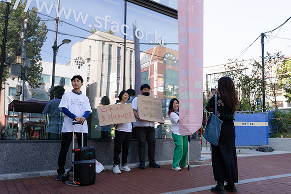 서울연극센터 건물 앞에서 ‘미투 이후 다시 말하기’ 퍼포먼스가 펼쳐지고 있다. 흰 티셔츠를 맞춰 입은 네 명의 사람이 연극센터 건물 앞에 일렬로 서 있다. 가장 왼쪽부터 각각 이동형 스피커, ‘미투 이후 좀 달라졌나요?’와 ‘반성폭력투쟁의 시간, 이제 우리 모두의 역사로’라는 손글씨가 쓰인 피켓을 들고 있고, 가장 오른쪽에 초록 바지를 입고 ‘세상아 너는 두려워해야 할 거야. 나는 생존자거든’이라는 문구가 적힌 긴 걸개 깃발을 든 최샘이 기획자가 밝은 얼굴로 웃고 있다. 길을 지나던 검은 옷의 한 행인이 네 명의 퍼포머를 마주보고 서 휴대폰으로 이들의 모습을 촬영하고 있다.