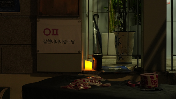 <재개발 옆 경로당>의 공연 사진. ‘갈현어버이경로당’이라는 흰 현판 옆으로 내부가 들여다보이는 철제 난간이 설치되어 있다.
난간 턱에는 불이 붙어 한쪽으로 기울어 타고 있는 제법 큰 양초와 손전등, 다소곳하게 포개진 <재개발 옆 경로당>의 포스터 여러 장이 나란히 놓여 있다.
난간 안쪽으로는 세 개의 화분과 두 개의 우산이 무심하게 놓여 있다.
난간 바깥쪽에는 검은 천이 씌워진 오브제 위에 ‘안전제일’이라고 쓰인 붉은 빗금이 들어간 테이프가 구겨져 있고,
그 옆에 온전한 모습의 안전제일 테이프와 주황색 종이테이프가 놓여 있다.