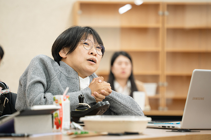 좌담 진행자 김지수. 짧은 커트 머리에 동그란 안경을 썼고, 흰색 셔츠 위에 회색 카디건을 입었다.
양손을 앞으로 모아 쥐고 조금 위쪽을 바라보며 말하고 있다.