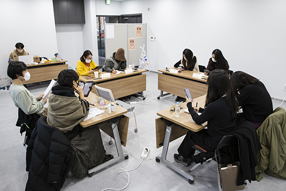 희곡결산수다회의 현장사진. 2명이 나란히 앉는 직사각형의 긴 책상 네 개가 정사각형 모양으로 배치되어 있다. 각 테이블에 두 명씩 총 여덟 명의 작가들이 둘러앉아 각자 태블릿이나 휴대폰, 인쇄물로 희곡을 보고 있다. 