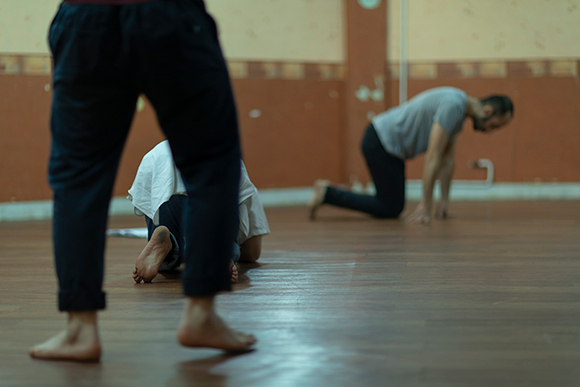 마룻바닥에서 훈련 중인 세 배우의 모습이다. 가장 앞쪽에 있는 배우는 뒷모습으로, 하체만 흐릿하게 보인다. 그 다리 사이로 바닥에 양 팔꿈치와 무릎을 댄 채 고개를 숙이고 있는 배우의 뒷모습이 선명하게 보이고, 멀리 똑같은 자세를 취한 배우의 전신이 흐릿하게 보인다.