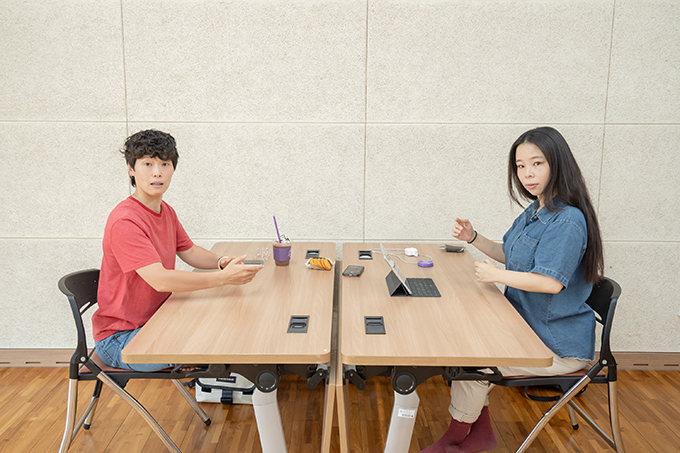 배우 성수연과 배우 권은혜가 책상을 마주 놓고 앉은 채 고개를 틀어 카메라를 응시하고 있다.
두 배우 모두 양팔의 팔꿈치를 구부리고 몸통에서 팔을 뗀 자세다.
배우 권은혜는 입을 살짝 벌리고 눈을 동그랗게 뜨고 있으며, 배우 성수연은 입을 다문 채 고개를 약간 기울였다.
