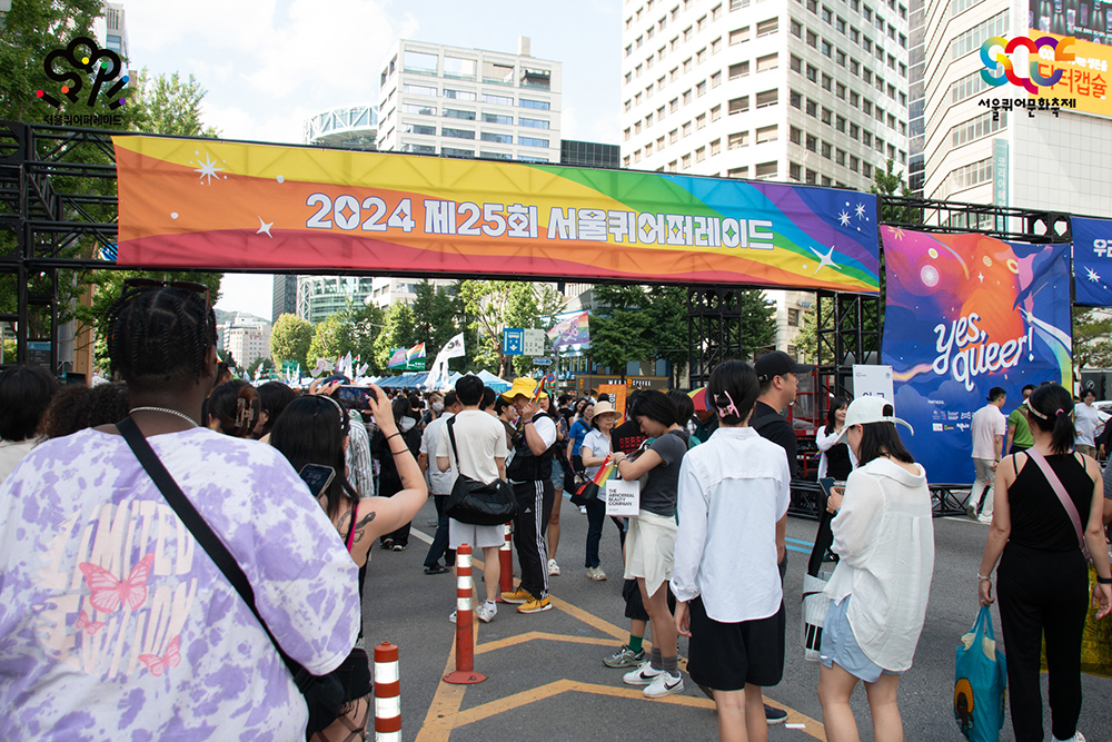 을지로입구역과 종각역 사이, 자동차 도로에 퀴어 퍼레이드에 참여한 사람들이 가득하다. 커다란 철제 구조물에는 무지개 배경에 ‘2024 제25회 서울퀴어퍼레이드’가 적힌 좌우로 긴 플래카드가 걸려 있고, 그 오른편에는 ‘yes, queer’가 적힌 정방형 플래카드가 걸려있다.