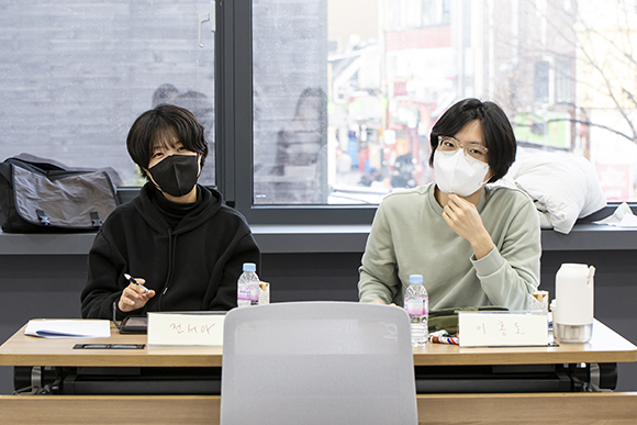 한 책상에 전서아 작가와 이홍도 작가가 나란히 앉아 정면을 바라보고 있다. 좌측에 앉은 전서아 작가는 검은 후드티와 검은 마스크를 썼고, 우측의 이홍도 작가는 밝은 회색의 맨투맨에 흰색 마스크를 착용했다. 두 사람의 뒤편에 있는 창 너머로 대학로의 풍경이 언뜻 보인다.