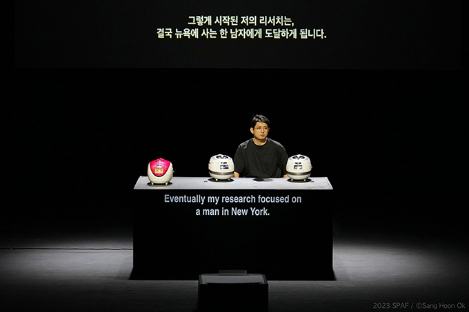 <쿠쿠>의 공연 사진. 검은 무대에 검은색 가로로 긴 직육면체 모양의 테이블이 하나 놓여있고, 그 위에 전기밥솥 세 개가 놓여있다.
제일 왼쪽에는 빨간색 구형 밥솥이 놓여있고, 우측 두 개는 흰색 신형 밥솥이다. 신형 밥솥 사이로 테이블 뒤에 앉은 구자하가 보인다.
검은 반소매 티셔츠를 입은 그는 얇은 뿔테 안경을 썼다.
무대 상단에는 ‘그렇게 시작된 저의 리서치는, 결국 뉴욕에 사는 한 남자에게 도달하게 됩니다’라는 한글 자막이 띄워져 있고, 테이블의 앞면에는 영문 자막이 영사되고 있다.