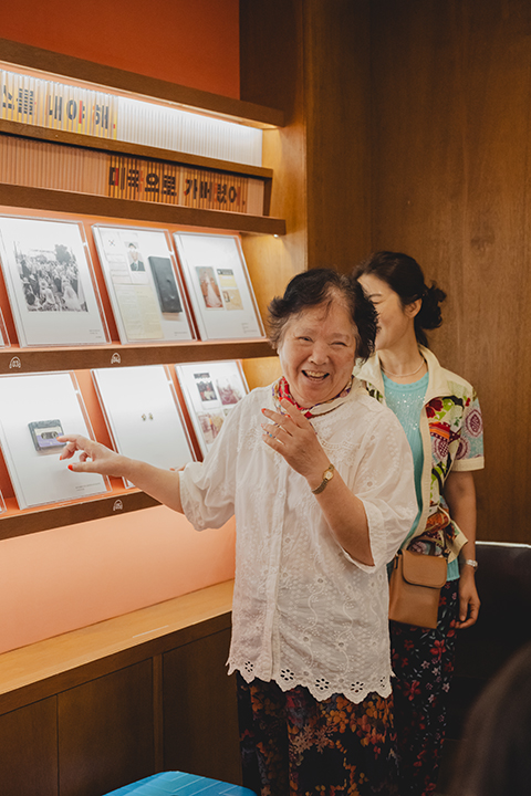 <오프 리밋> 현장 사진. 미군 청년 ‘영철’과의 사랑 이야기를 들려주는 김숙자 할머니. 
            이 공간은 기지촌여성평화박물관의 한 전시실로, 이곳에는 할머니들이 당시 사용했던 사적인 물건들이 전시되어 있다. 
            전시 벽면에 쓰인 “미국으로 가버렸어”라는 문장이 눈에 띈다. 
            김숙자 할머니는 ‘영철’이 녹음해줬던 노래가 담겨 있는 카세트 테이프를 가리키며 웃고 있다.