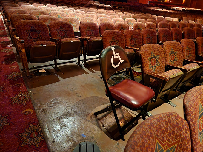 극장 객석의 중간쯤, 고정된 좌석들 사이에, 통로 옆 넓은 공간이 비어 있고, 
                휠체어 이용인의 이미지가 그려진 접이식 의자가 하나 놓여 있다.