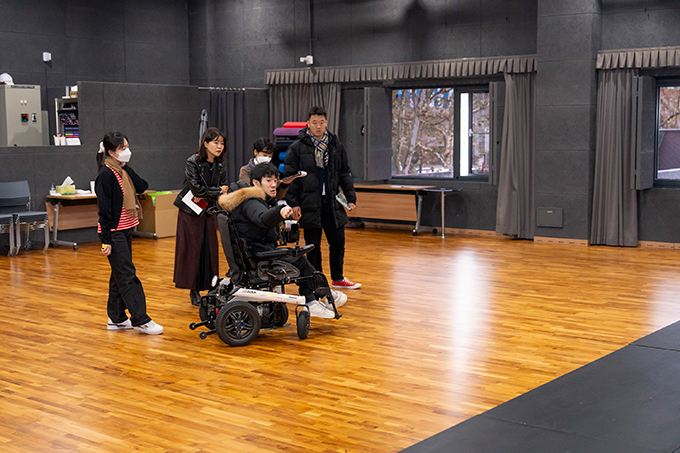 3층 스튜디오의 접근성 점검 현장. 휠체어를 이용하는 퍼실리테이터와 함께 조를 이룬 네 명의 참가자들이 모여 있다. 
                    퍼실리테이터가 오른손을 들어 경사로가 없는 덧마루 무대를 가리키는 모습이다.