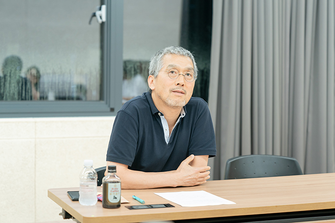 연출가 강량원. 동그란 안경을 쓰고 칼라가 있는 남색 반소매 티셔츠를 입었다.
흰 머리와 검은 머리가 섞여 머리가 은빛으로 보인다. 먼 곳으로 시선을 보내고 있다.