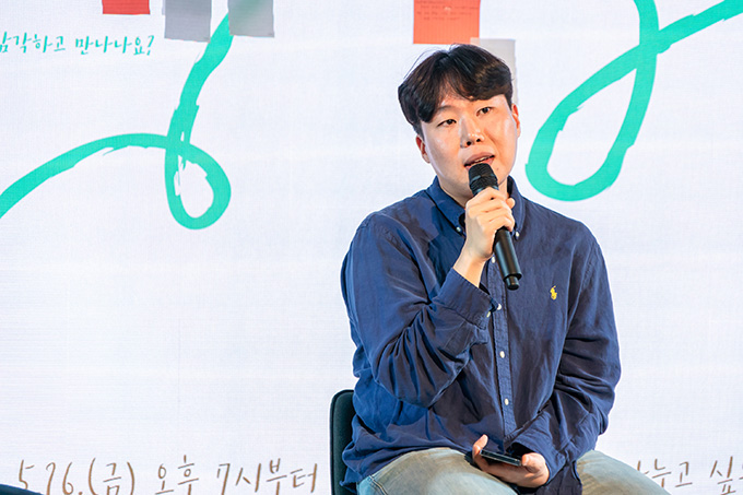 작가 배해률. 살짝 웨이브가 있는 앞머리. 푸른색 셔츠에 옅은 청바지를 입고 있다. 왼손에는 스마트폰을, 오른손에는 마이크를 들고 이야기하는 중이다.
