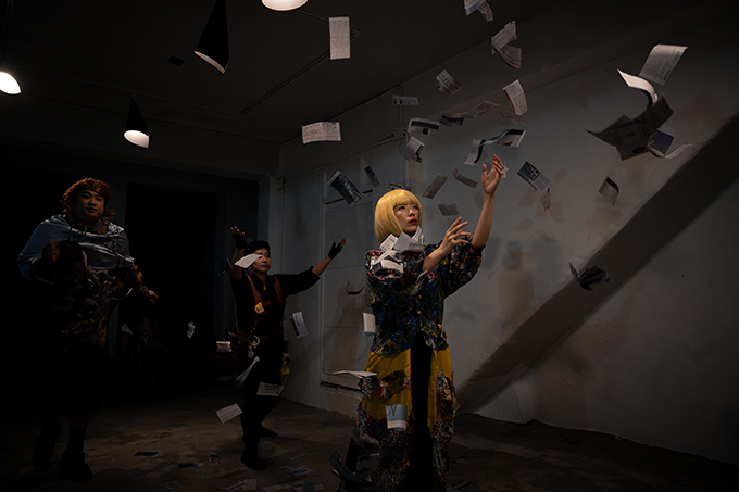 <쇼 팔다>의 공연 사진. 어두운 공간, 천장에는 종이로 만든 갓을 씌운 전구 네 개가 걸려 있다. 
            공간에 손바닥보다 조금 더 큰 크기의 직사각형으로 잘린 신문지들이 휘날리는데 세 인물이 두 팔을 들고 그 사이를 걸어간다. 
            맨 앞쪽에 선 인물은 노란 단발머리에 앞머리를 내리고, 화려한 무늬의 재킷과, 재킷의 무늬와 유사한 천들을 듬성듬성 덧댄 노란 치마를 입었다. 
            그 뒤로 손가락장갑을 끼고 검은 옷을 입은 짧은 머리의 인물이 보이는데, 작업 조끼에는 줄자를, 허리춤에는 테이프와 케이블타이 등을 매달고 있다. 
            그 옆의 인물은 갈색의 곱슬머리 가발을 쓰고, 은색 망토를 두르고 있지만 어두워 의상이 잘 보이지 않는다. 