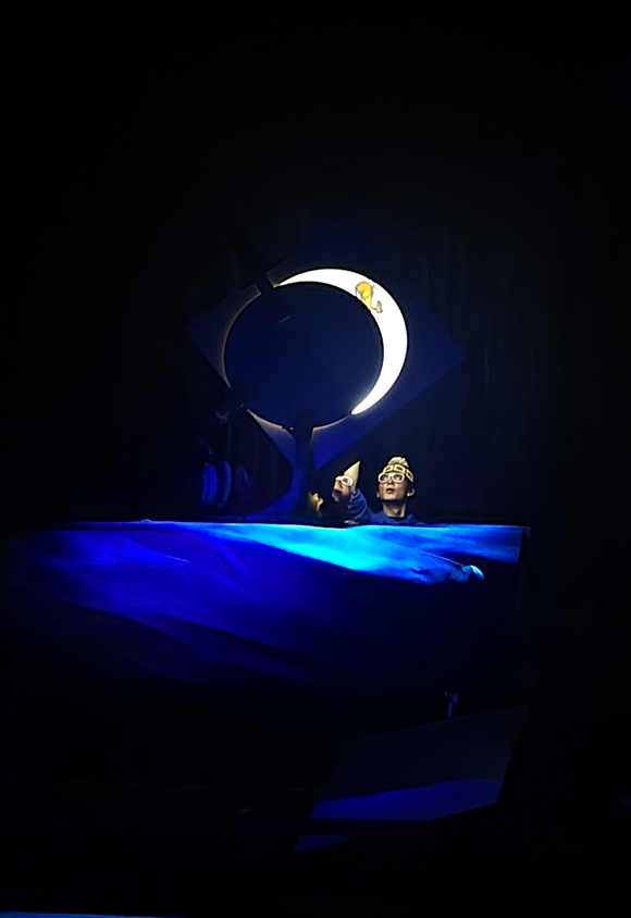 <해와 달에 관한 오래된 기억>의 공연사진이다. 바다처럼 보이는 푸른 천이 배우들의 어깨 높이까지 펼쳐져 있고, 배우들은 달 모양의 조명을 응시하고 있다. 위 아래가 뒤집힌 초승달 모양 가운데에 노란 고래 한 마리가 있다. 