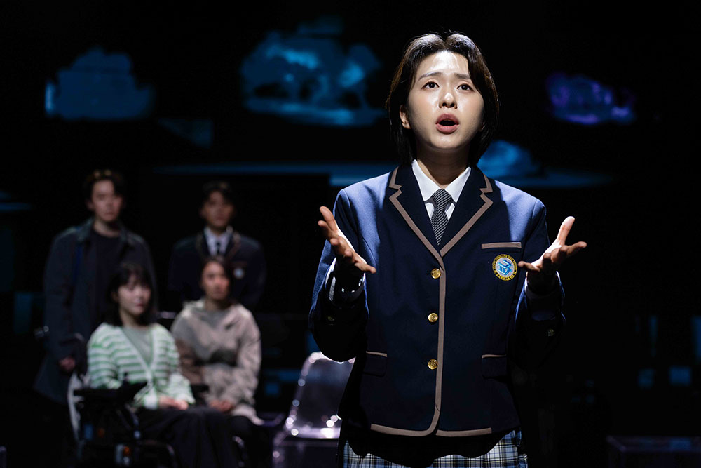 <천 개의 파랑>의 공연 사진. 남색 교복을 단정히 입은 단발머리 연재가 양손을 활짝 펼쳐 가슴께에 들고 정면을 바라보며 무언가 말하고 있다. 그 뒤편으로 그의 가족들이 각자 앉거나 서서 말하는 연재를 응시한다.