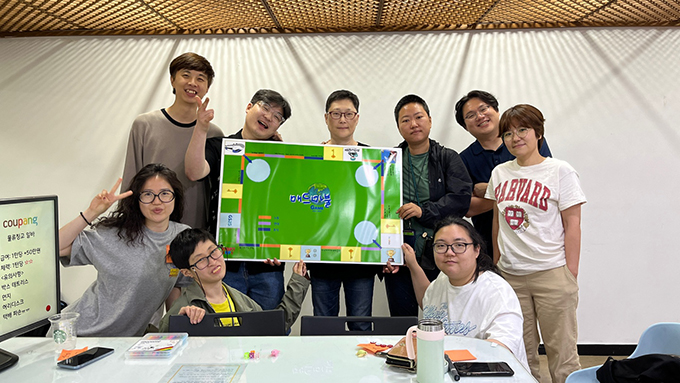 미친회사 프로젝트의 구성원들이 모여 기념사진을 찍는다. 커다란 ‘매드마불’ 게임판을 다 함께 들고 카메라를 보며 웃음 짓고 있다.