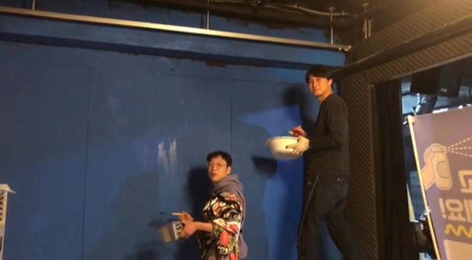 페인트를 담은 그릇과 붓을 든 두 사람이 파란색 벽 앞에 서서 고개를 돌려 정면을 바라본다.
한 사람은 무언가를 밟고 올라선 듯, 다른 한 사람보다 훨씬 높은 곳에 서있다. 그들이 칠하는 벽의 오른편에 방음스펀지가 붙은 벽이 보인다.