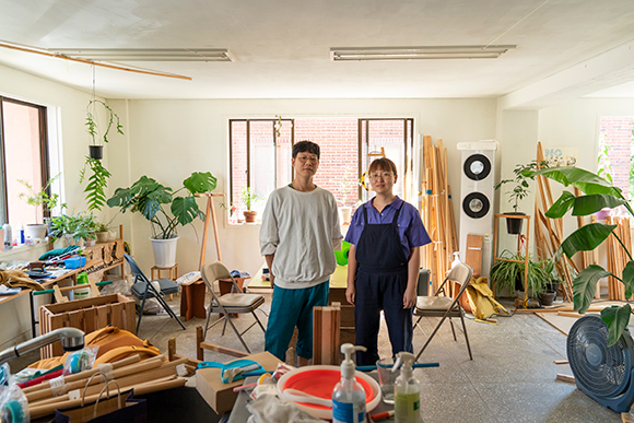 이두호, 신원정 두 사람이 작업실 공간에 나란히 서 있다. 식물들과 목재들, 각종 오브제들이 공간 여기저기 쌓여 있다.