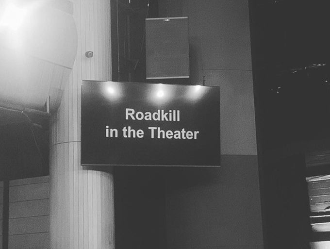 극장 객석 앞에 설치된 자막용 모니터. 흑백사진이다. 검은 바탕의 화면에 흰색으로 “Roadkill in the Theater”라고 쓰여 있다. 모니터 위쪽으로 스피커가 매달려 있으며,
화면에는 밝은 조명 세 개가 반사되어 빛난다.