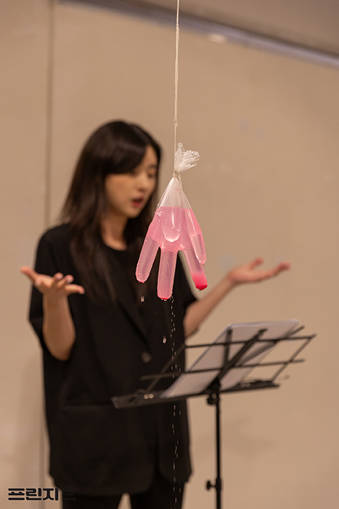 <녹로수> 퍼포먼스 사진. 천장에서부터 늘어뜨린 실에 묶인 비닐장갑에 핑크색 액체가 담겨 있다. 장갑에 작은 구멍이 나 물이 방울방울 떨어진다. 
            그 뒤로 보면대에 대본을 놓고 연기하는 퍼포머의 모습이 흐릿하게 보인다. 그는 가슴까지 오는 생머리에 한쪽만 앞머리를 내렸고, 검은 반소매 재킷과 검은 티, 검은 바지를 입었다.