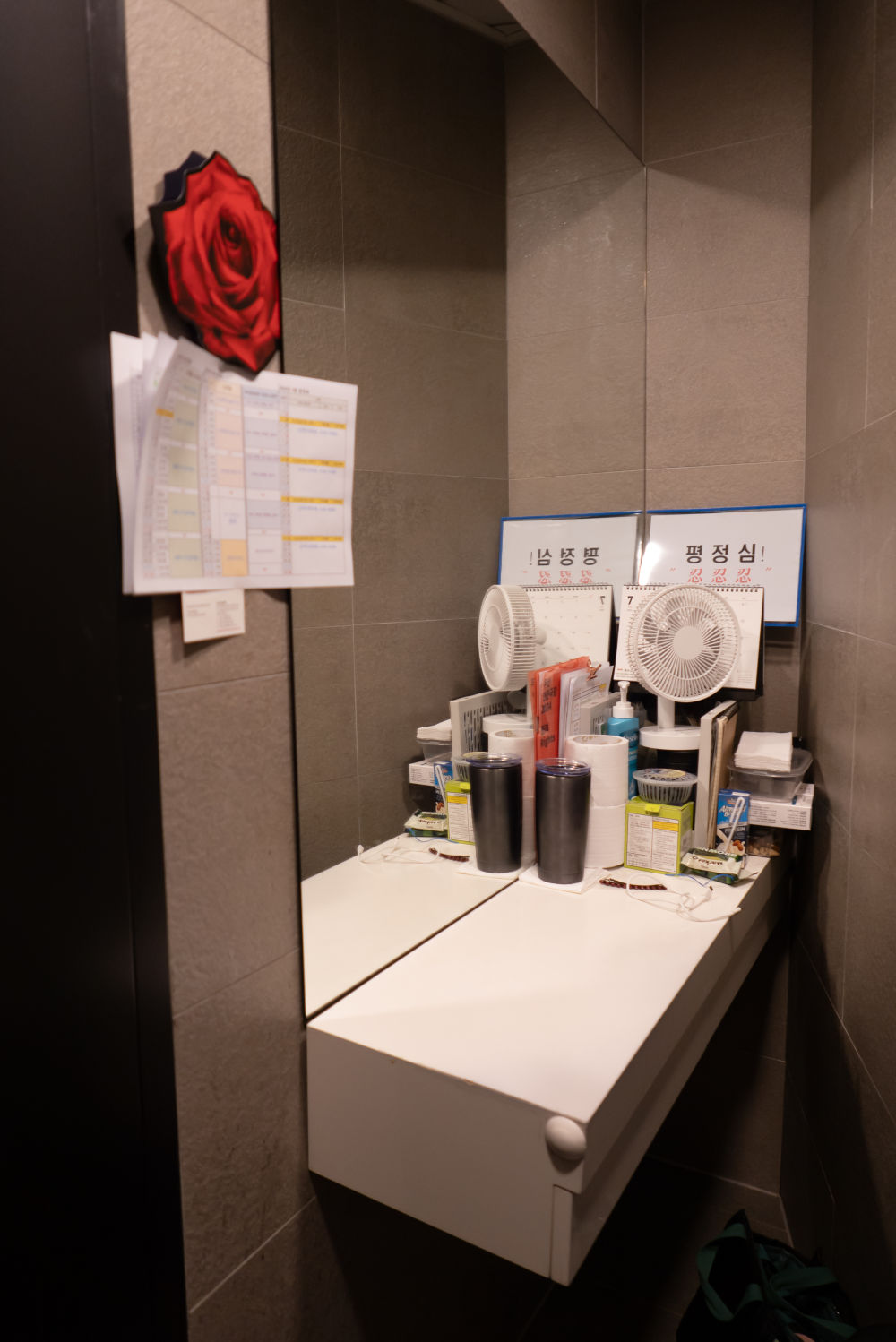 타일 벽에 화장대와 거울이 설치된 좁은 공간을 촬영한 사진. 화장대의 오른쪽에는 텀블러와 휴지, 약간의 간식, 선풍기, 책상달력 등이 단정하게 놓여있다. 달력 뒤에는 평정심과 참을 인 세 개를 한 페이지에 인쇄하여 코팅한 종이가 놓여있다. 거울 왼쪽 벽에는 일정표와 붉은 장미 그림이 붙어있다.