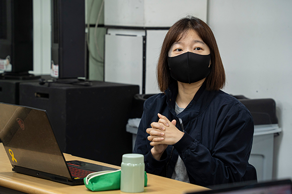 참여자 박하늘. 남색 재킷을 입고 검은색 마스크를 썼다. 두 손을 모아 깍지 낀 채 이야기하고 있으며, 책상에는 노트북이 펼쳐져 있다.