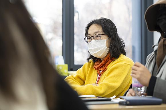 작가 일요. 어깨를 가볍게 덮는 머리는 파마기가 남아있고, 검고 둥그런 안경을 썼다. 노란 가디건에 주황색 손뜨개 목도리를 하고 있다. 