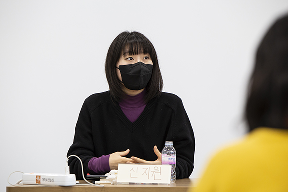 작가 신지원. 어깨를 살짝 덮는 단발에 앞머리를 내리고 검은 마스크를 썼다. 보라색 목폴라 위에 검은 가디건 차림이다. 
