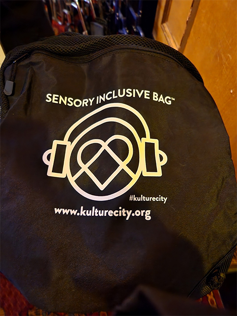 검은색 가방에 “SENSORY INCLUSIVE BAG”이라는 글씨와 함께, 
                동그라미와 하트, 헤드셋 등을 연상시키는 이미지가 그려져 있다.