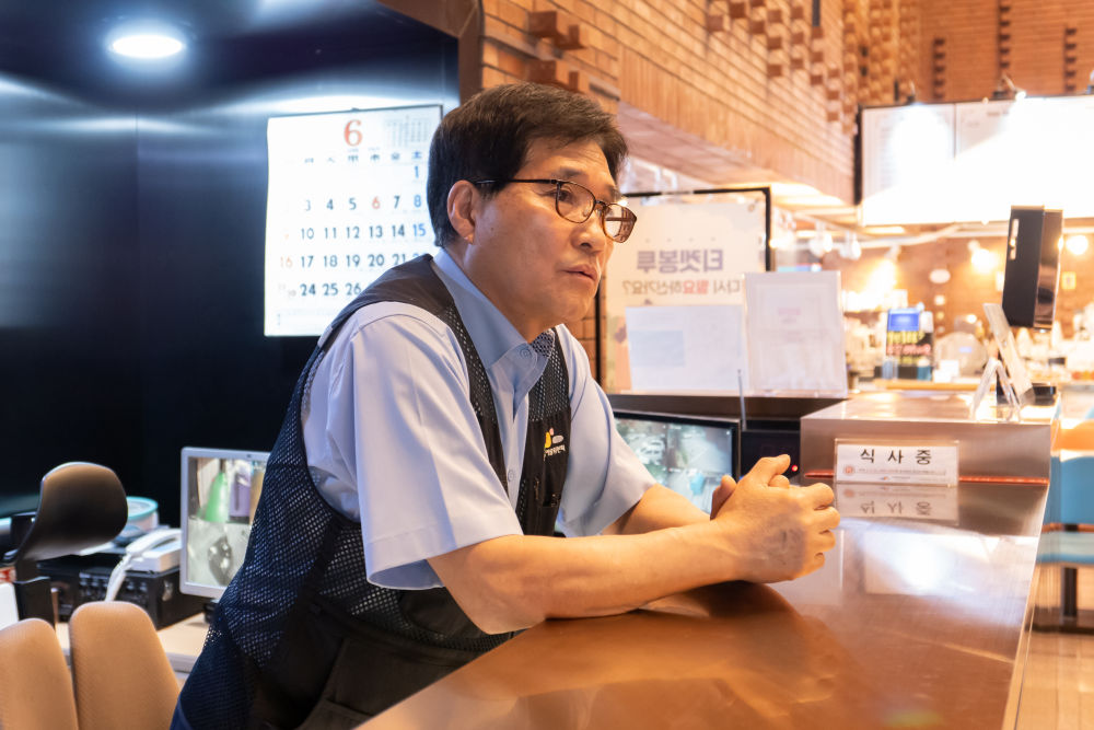 아르코 예술극장 로비. 김명식이 안내 데스크에 두 팔을 걸친채 서서 이야기한다. 그는 하늘색 반소매 셔츠 위에 한국문화예술위원회 로고가 있는 작업조끼를 걸치고 안경을 썼다.