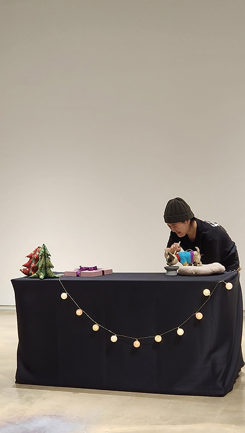 <칼럼버스 프로젝트> 중 <선물>의 공연 사진. 검은 천이 덮인 허리 높이 정도 되는 테이블의 앞면에 알전구가 늘어져 있다. 
            테이블 위에는 왼편에 빨간색, 초록색의 작은 트리 인형 두 개와 보라색으로 포장된 선물이 놓여 있다. 
            오른편에는 방석 위의 강아지 인형이 밥을 먹는 듯 밥그릇에 고개를 들이밀고 있는데, 
            한 손으로 인형을 조종하는 퍼포머가 웃음 띤 얼굴로 그 모습을 바라보고 있다.  