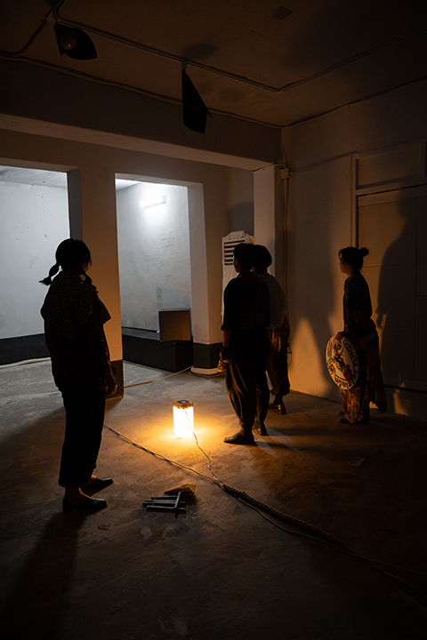 <쇼 팔다>의 공연 사진. 벽면에는 흰 페인트가, 바닥에는 시멘트가 칠해진 공간이 몇 개의 기둥을 기준으로 둘로 나뉘어 있다. 
            한쪽 공간에 네 명의 배우가 서 있는데, 이곳을 밝히고 있는 것은 바닥에 놓인 주황색 램프뿐이다. 
            이들은 모두 기둥 너머 불이 밝혀진 또 다른 공간을 바라보고 있다. 램프에 비친 배우들의 그림자가 벽면을 가득 채우고 있다. 