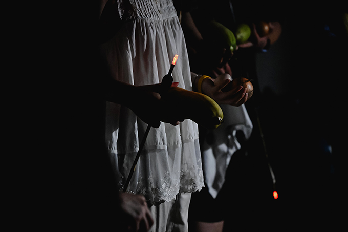 <정전의 밤> 공연 사진. 어둠 속에 일렬로 늘어선 사람들의 상반신이 보인다. 이들은 양손에 양파, 애호박, 달걀 등과 화살을 들고 있다.