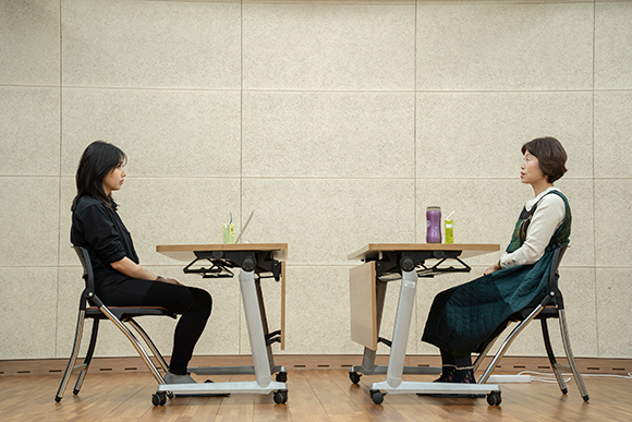 배우 박은호와 배우 이미경이 각자의 책상 앞에 앉아 서로를 마주 보고 있다. 두 사람의 배경으로, 밝은 베이지색 벽에 나무 바닥의 공간이 보인다.