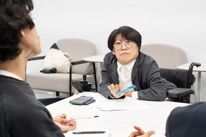 좌담 진행자 김지수. 하얀색 셔츠의 단추를 목까지 채우고 그 위에 짙은 회색의 재킷을 입고 있다. 
            짧은 커트 머리에 동그란 안경을 썼다. 오른손에 볼펜을 들고 있다.