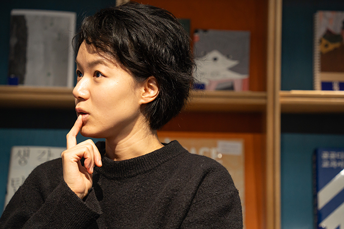 작가 강수연. 웨이브가 있는 커트 머리. 검은색 라운드 넥의 긴소매 니트를 입었다.
오른손 검지를 입가에 대고 골똘히 생각에 잠긴 모습이다.