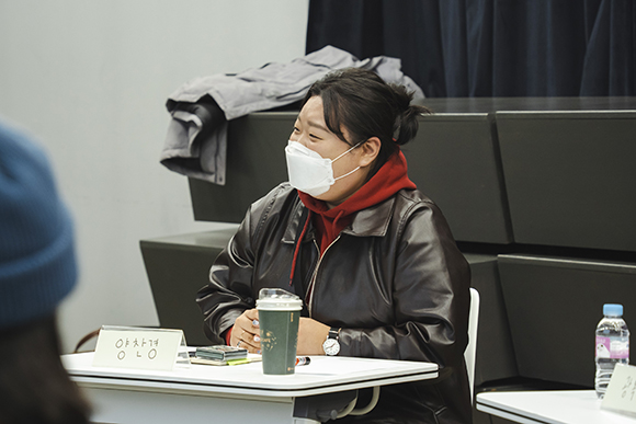 작가 문서희. 단발 머리를 가볍게 묶고, 빨간 후드티에 검은 가죽자켓을 걸쳤다. 양손을 가볍게 깍지 끼고 측면을 바라보며 가볍게 웃음짓고 있다. 책상 위에 놓인 이름표에 ‘양찬경’이라고 쓰여 있다. 